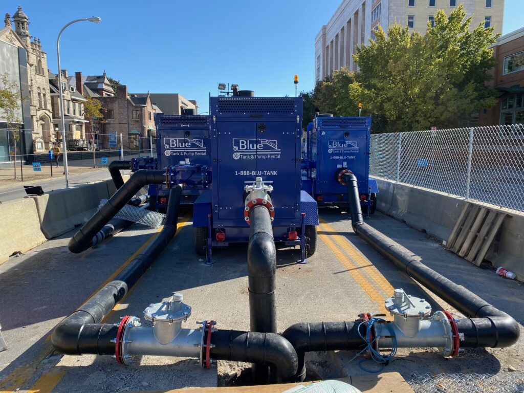 A sewer bypass setup on a city street, with large blue pumping units connected to thick black hoses that run along the ground.